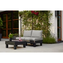 Modern, szürke párnázott elemekből álló balkon kertibútor szett az erkélyen, mellette fekete kávézóasztal, virágcserép és üveg víz, zöld növényekkel körülvéve, hangulatos környezetben.