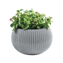 Felakasztható, szürke műanyag kaspó horgolt mintázattal, benne zöld levelű növényekkel és rózsás virágokkal. Modern, időjárásálló kialakítás, ideális belső és kültéri használatra.