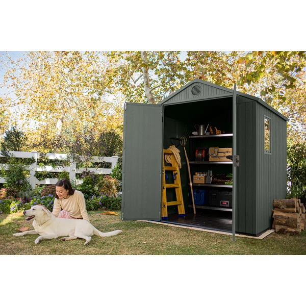 Zöld, műanyag kerti ház a nyitott ajtóval, mögötte különböző szerszámok és tárolódobozok láthatók. Előtte egy nő ül a fűben, kezében egy fehér kutyával a barátságos kertben, napsütéses időben.