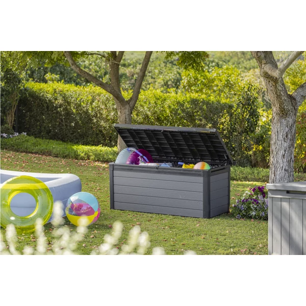 Kültéren álló, sötétszürke műanyag kerti tároló láda, nyitott fedéllel, amelyben különböző színes labdák láthatók. A láda körül zöld fű és virágok találhatók, napfényes környezetben.