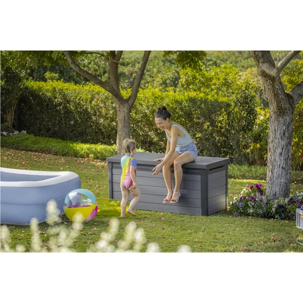 Kerti környezetben, kinyitott fedelű CORTINA műanyag tárolódoboz, mellette egy gyermek játékos tevékenységet végez, míg egy felnőtt mosolyogva interakcióba lép vele. A doboz időjárásálló kompozitból készült, szürke színű.