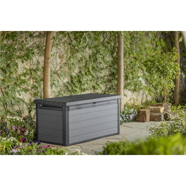 Szürke műanyag kerti tárolóláda a Cortina sorozatból, kültéri használatra tervezve, hidraulikus zárható fedéllel és UV-védelemmel ellátva, virágokkal és fákkal díszített kertben elhelyezve.