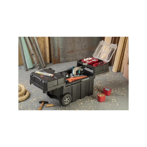 Mobil KETER 'MASTERLOADER' szerszámosláda, fekete színben, gumi kerekekkel és puha markolatú fogantyúval, vízszintesen nyitható tetővel. Kivehető rekeszekkel és biztonságos zárral ellátva, barkács környezetben, fűrészporral borítva.