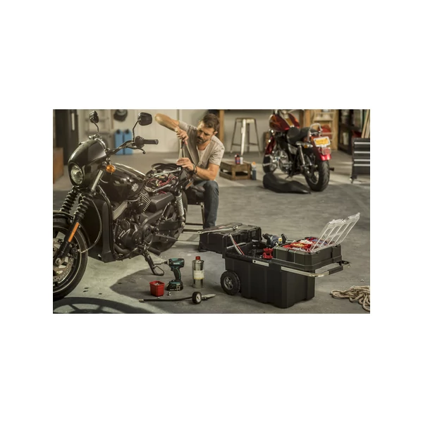 A képen egy férfi éppen egy motor javításával foglalkozik egy garázsban, mellette a földön egy KETER 'Masterloader' szerszámosláda található, amely kerekeken gurul és telis-tele van szerszámokkal és kivehető rekeszekkel.