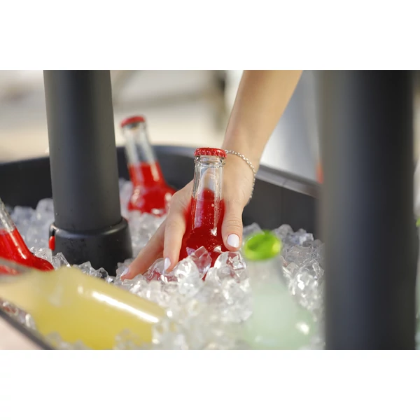 Kéz, amely egy üveg vörös üdítőt emel ki egy jégágyon fekvő műanyag italhűtőből, szabadban készült a nyári napfényben. A környezet alkalmas baráti összejövetelhez vagy családi találkozóhoz.