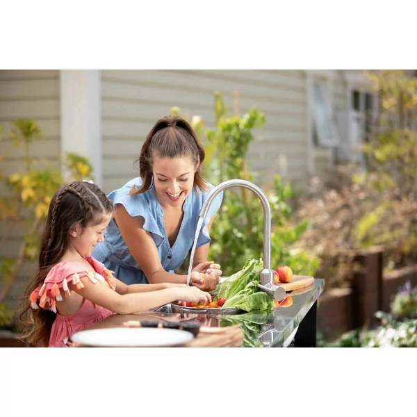 Kerti grillasztalon, a Unity Chef mosogató mellett egy felnőtt nő és egy lány gyermek friss zöldségeket készítenek elő, vidám környezetben, zöld növények között, napsütéses időben.