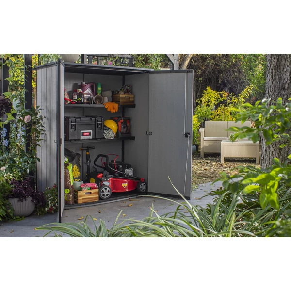 Szürke műanyag tároló, nyitott ajtókkal, polcokkal és különböző tárgyakkal tele. Kertben található, zöld növényzet veszi körül. Praktikus tárolási megoldás kültéri használatra.