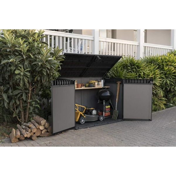 Szürke, műanyag Premier Jumbo kerti tároló, nyitott állapotban, benne szerszámok és kerti kiegészítők láthatók. Az eszközök egy polcon helyezkednek el, mögötte zöld növények és tégla járda található, kültéri környezetben.