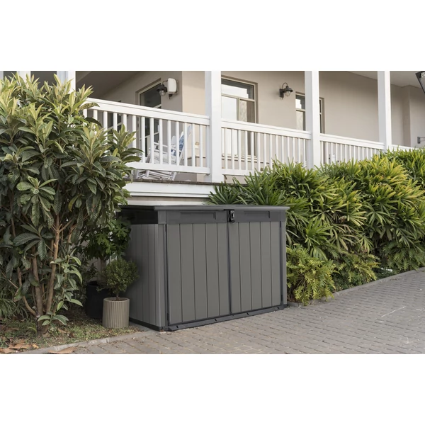 Prémium, szürke műanyag kerti tároló garázs előtt, zárt állapotban. A tároló a kerti kiegészítők védelmét szolgálja, időjárásálló és karbantartást nem igényel. Zöld növények között helyezkedik el.