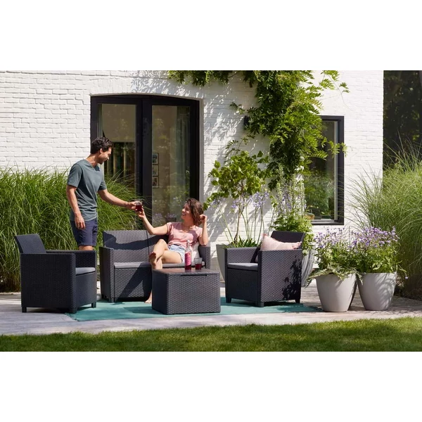 Kertben elhelyezkedő Claire műrattan kerti bútor szett, párnátartós asztallal. Két fotel és egy kávéasztal látható, kényelmes párnákkal, kedvező, napfényes környezetben, zöld fű és virágok között.