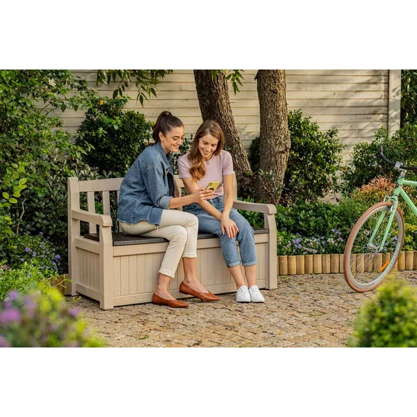 Két nő ül egy EDEN GARDEN műanyag kertipadon a szabadban, közvetlenül egy zöld, virágos környezetben. Az egyikük telefonozik, miközben a másik figyeli. A pad stílusos és funkcionális, tárolóként is funkcionál.