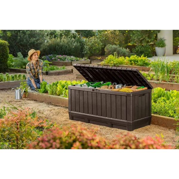 Kertben elhelyezett sötétbarna műanyag tárolódoboz, nyitott tetővel, benne kerti szerszámok és eszközök láthatók. A háttérben zöldségeskert és virágoskert található, a tároló ülőhelyként is funkcionál.