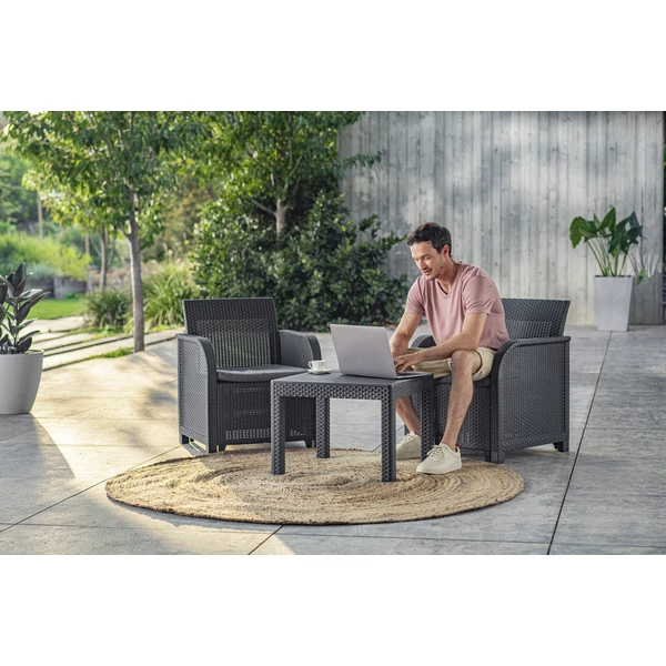 Két fekete műrattan fotelből és egy asztalból álló Rosalie erkély szett, természetes környezetben, burkolt teraszon, szőnyeggel, a mellettük ülő férfi laptopozik, kávéval. Kényelmes kivitel, puha párnákkal.